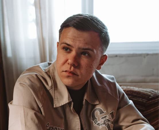 A portrait of a young woman with curly reddish-brown hair and bangs, wearing a red sweater against a solid yellow/orange background.